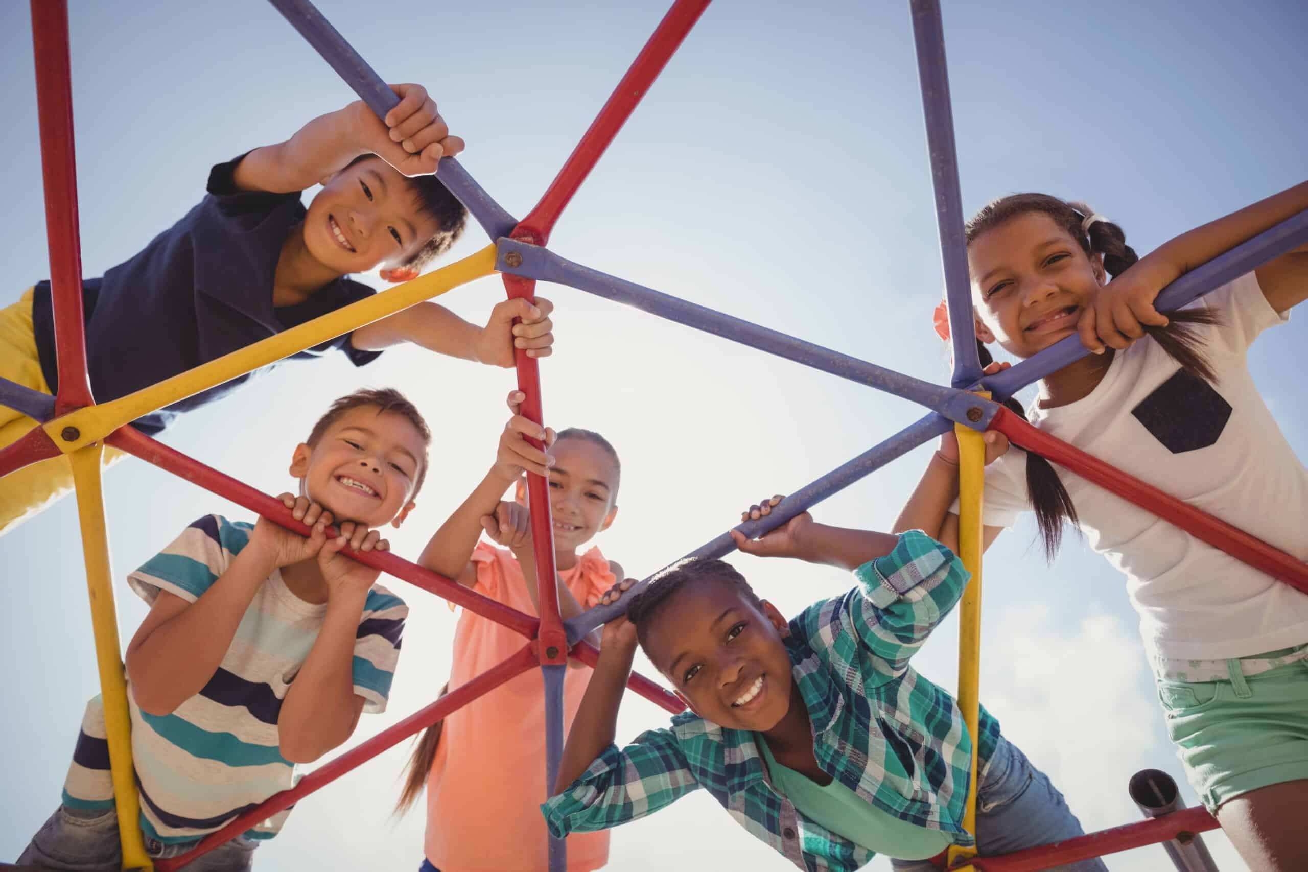 Five children smile and play on a playground climbing frame, looking down through the bars at the camera on a sunny day.
