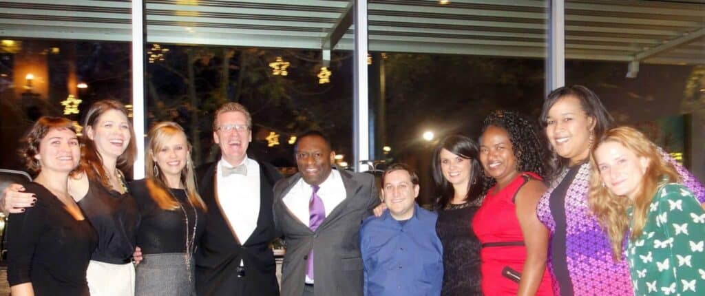 Ten people in formal and semi-formal outfits stand together indoors, smiling at the camera.