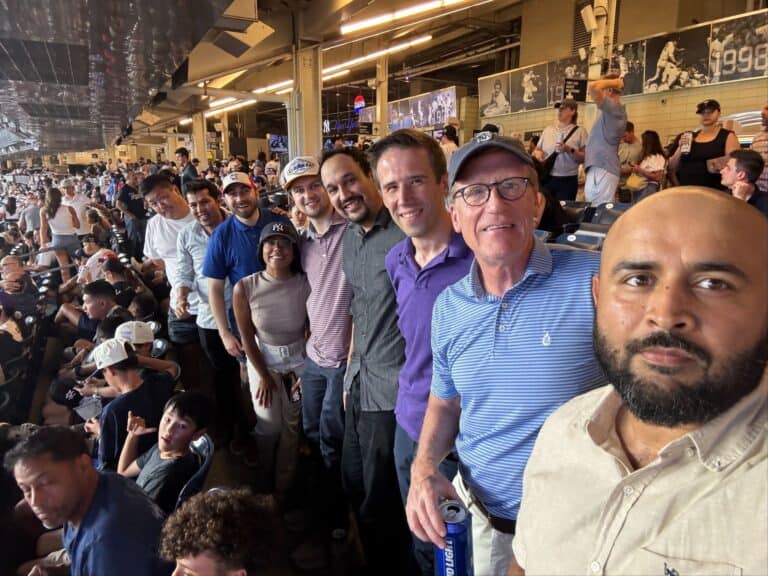 Ten adults stand in a row, smiling at a crowded stadium with many spectators filling the background.