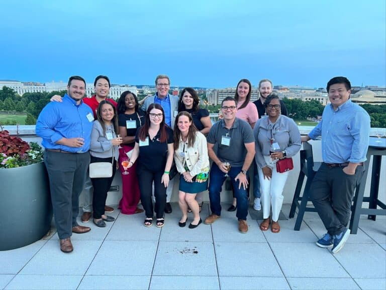 Thirteen smiling adults pose together on a rooftop patio, with city buildings and trees visible in the background.