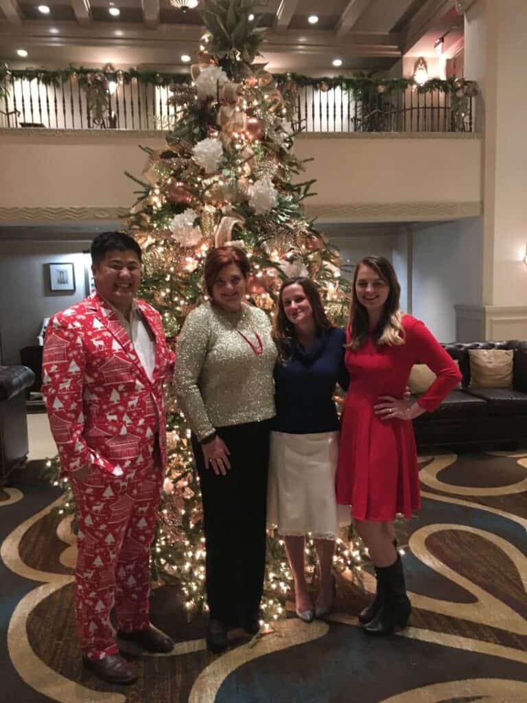 Four people in festive attire pose for a photo in front of a Christmas tree at a party