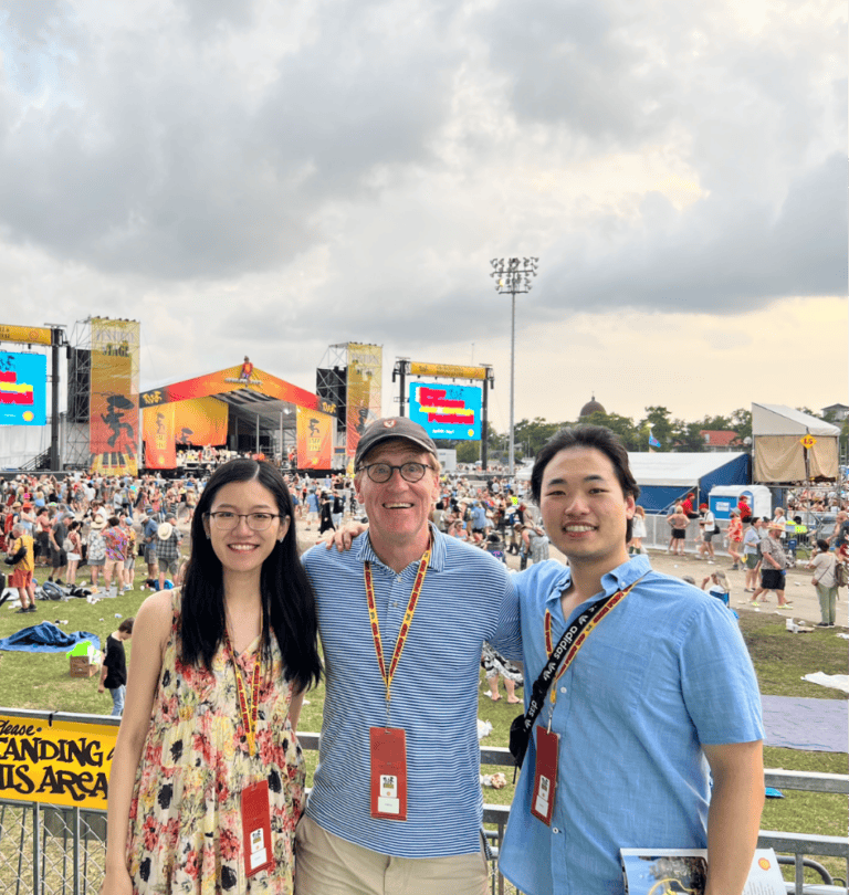 Three people smile while standing in front of a large outdoor festival stage, with a crowd and cloudy sky behind them.