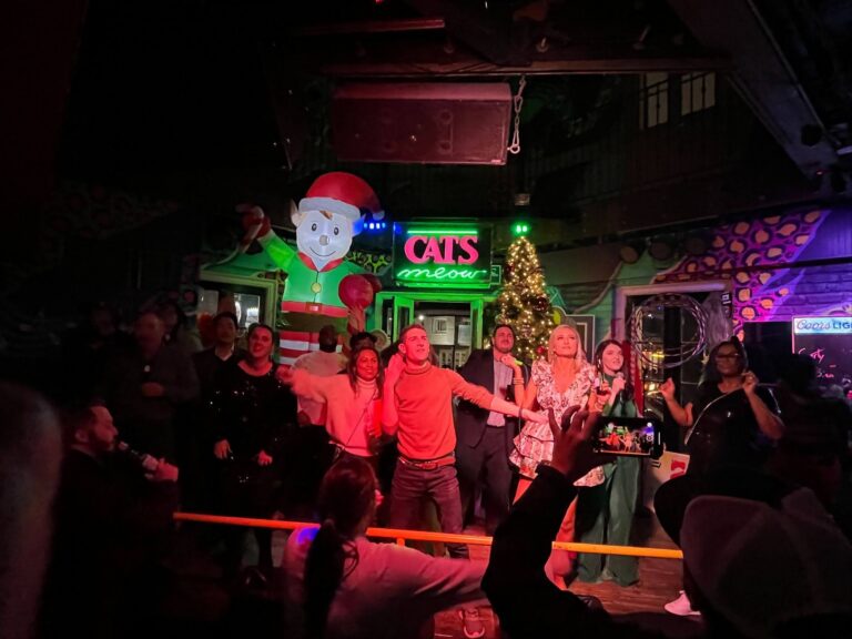 People sing and pose on stage at a festive bar with Christmas decorations, including a Santa, a tree, and glowing 