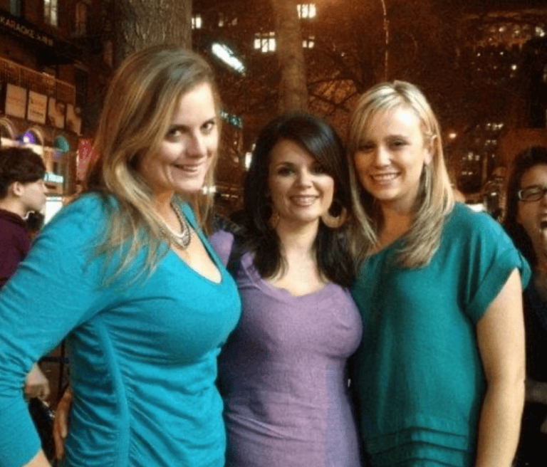 Three women pose outdoors at night; two wear teal tops, one wears a lavender dress, with city lights twinkling in the background.