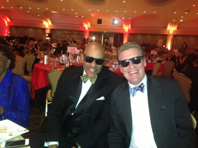 Two men in suits and bow ties, wearing sunglasses, smile while seated at a formal event with round tables and red lighting.