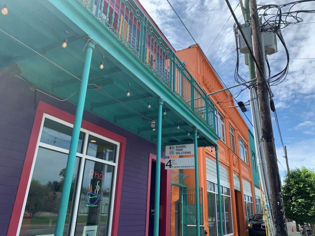 A vibrant building with a teal balcony, purple and orange walls, a pho restaurant sign, and power lines beneath a partly cloudy sky.
