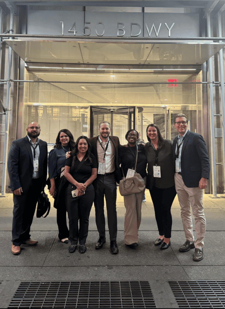 Seven people smile and pose for a group photo at night in front of the glass entrance at 1450 Broadway, New York.