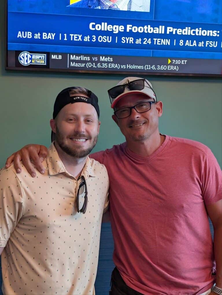 Two men stand close together, smiling at the camera, with a TV in the background showing sports news.