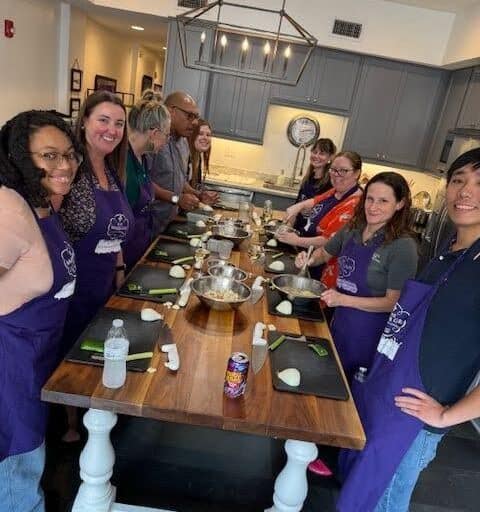 People of various backgrounds in purple aprons gather around a wooden table preparing to cook with fresh ingredients.