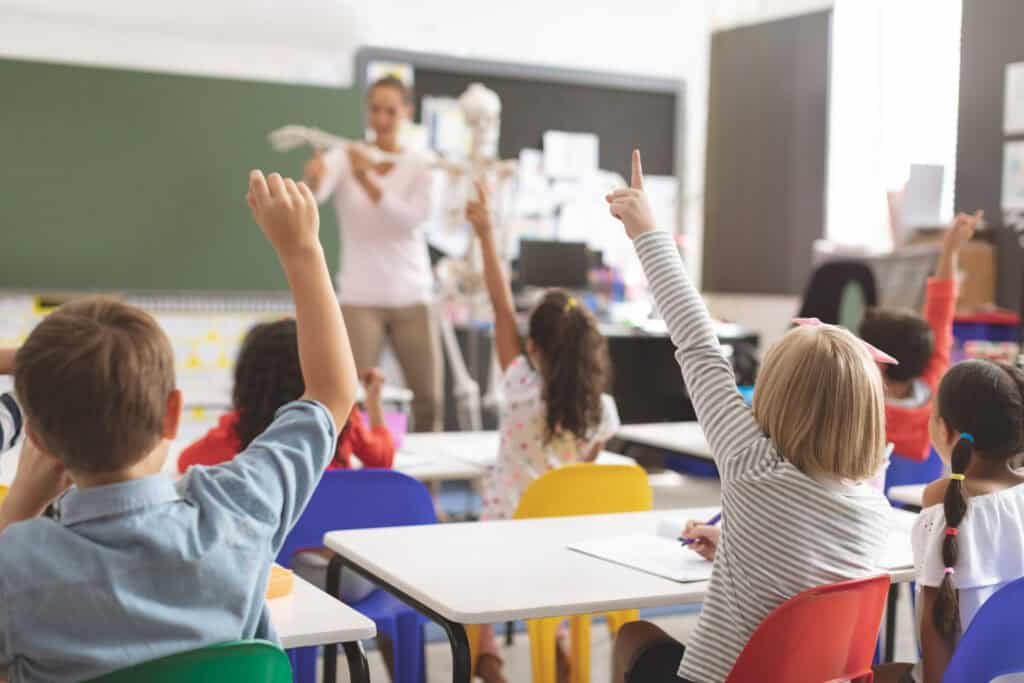 Students seated at desks raise their hands as a teacher explains accounting near a chalkboard and a skeleton model in the classroom.