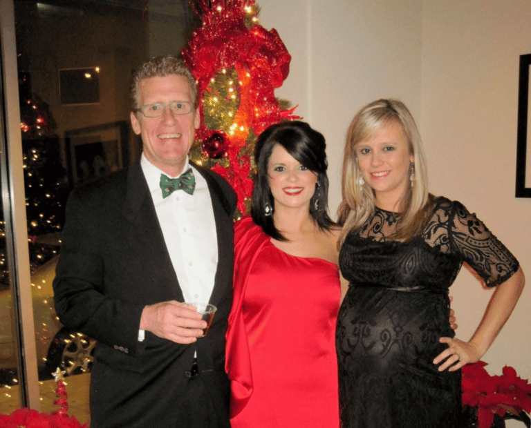 Three people dressed in formal, festive clothing stand indoors by a decorated Christmas tree, with red poinsettias nearby.