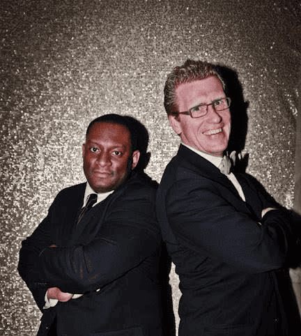 Two men in dark suits stand back to back with arms crossed, posing before a glittery, textured gold backdrop.