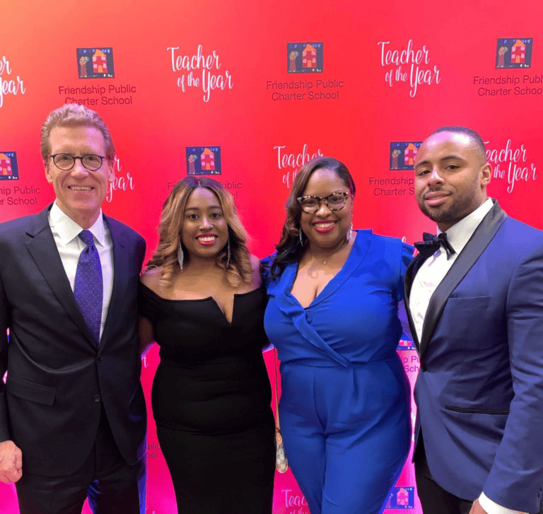 Four adults in formal wear stand before a red backdrop featuring “Teacher of the Year” and “Friendship Public Charter School” logos.