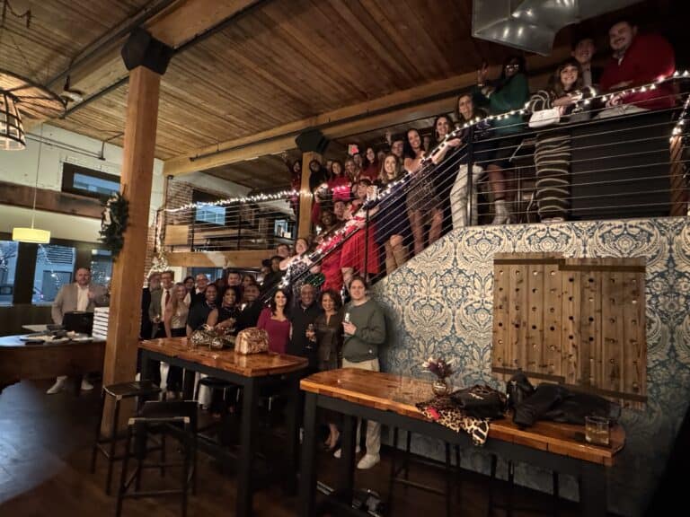 A large group poses for a photo on a multi-level indoor balcony decorated with string lights.