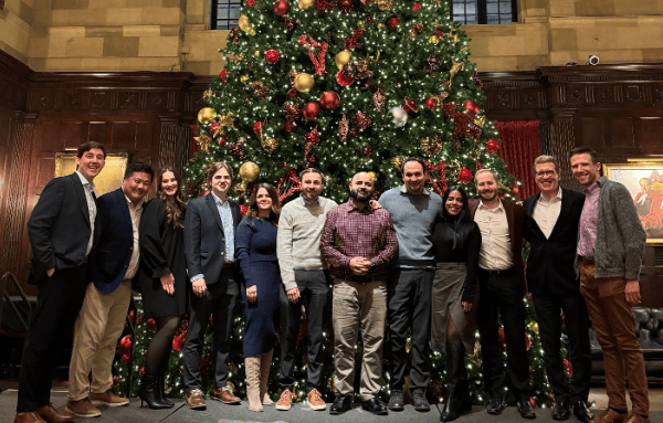 Twelve people stand before a tall, decorated Christmas tree in a grand room with wood paneling and high ceilings.