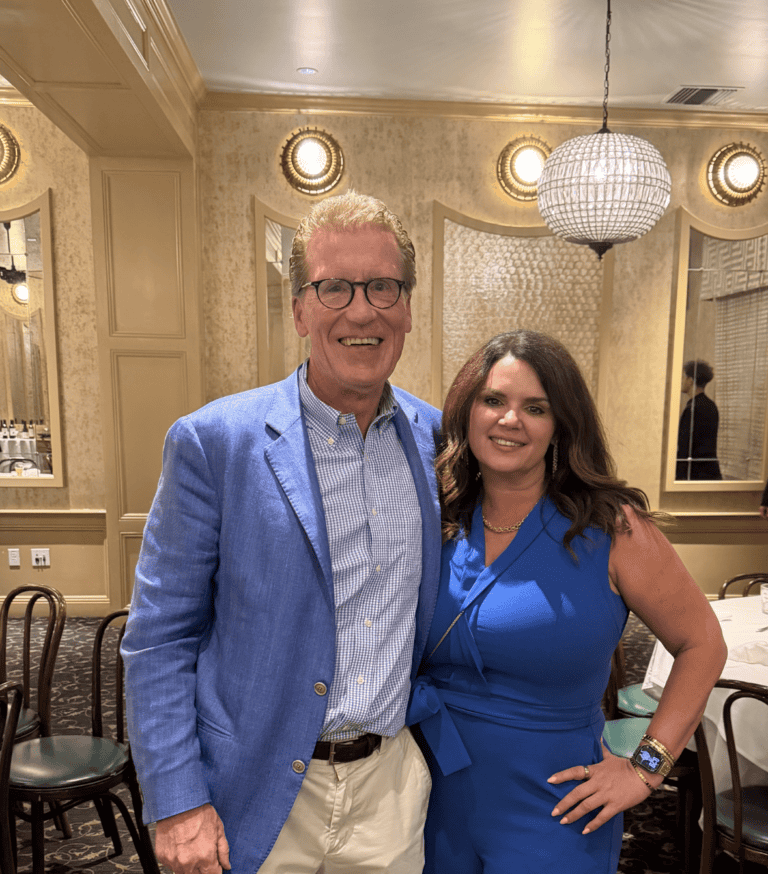 A man in a blue blazer and a woman in a blue jumpsuit pose together in an elegantly decorated restaurant.