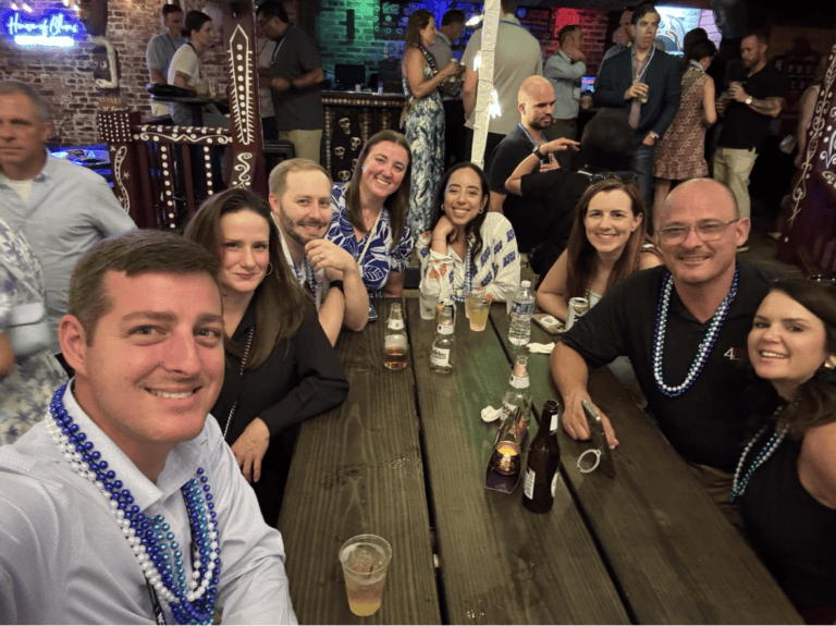People smile at the camera while sitting at a wooden table with drinks in a lively, decorated indoor setting.